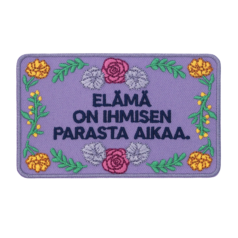Brodeerattu kangasmerkki, kukkia, teksti: Elämä on ihmisen parasta aikaa.