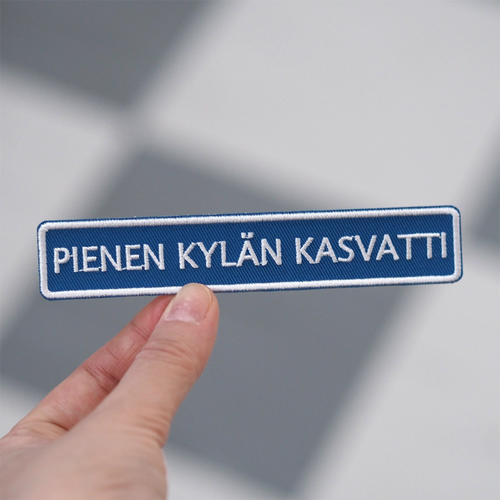 Brodeerattu haalarimerkki, tiekyltti, teksti: Pienen kylän kasvatti. Merkki henkilön kädessä.
