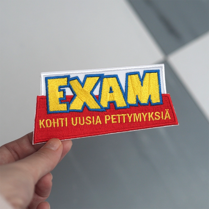 Brodeerattu haalarimerkki, teksti: Exam - kohti uusia pettymyksiä. Merkki henkilön kädessä.