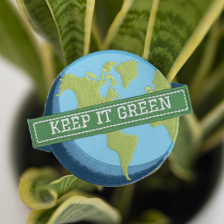 Brodeerattu maapallo-aiheinen haalarimerkki, teksti: Keep it green. Taustalla viherkasvi.