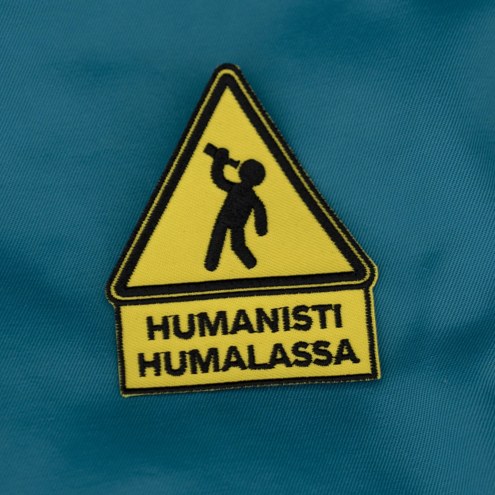 Brodeerattu haalarimerkki, liikennemerkki, teksti: Humanisti humalassa. Taustalla opiskelijahaalarit.