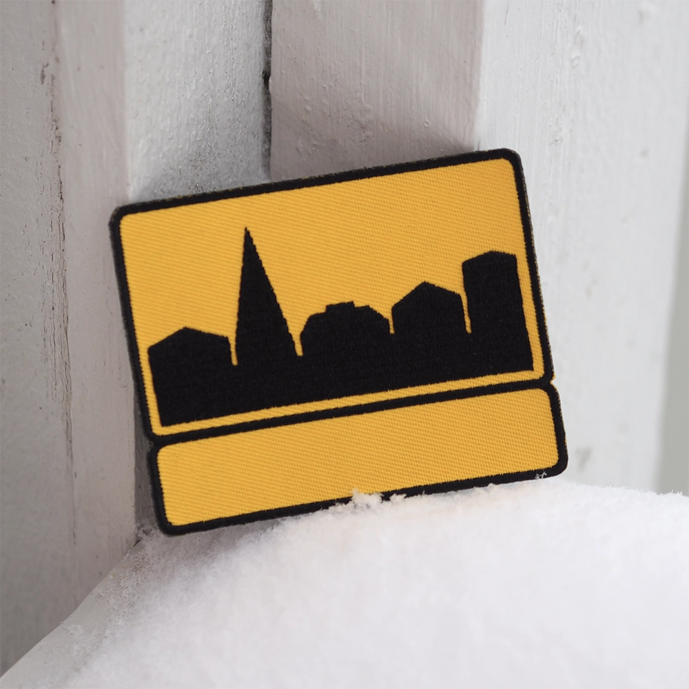 Brodeerattu liikennemerkki-aiheinen haalarimerkki, keltainen. Taustalla lunta ja seinä.