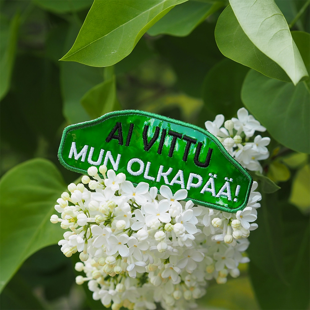 Brodeerattu hologrammihaalarimerkki, teksti: Ai vittu mun olkapää! Taustalla kukkia.