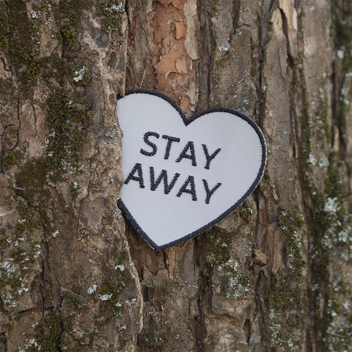 Brodeerattu heijastin haalarimerkki, sydämen muotoinen, teksti: Stay away. Taustalla puun runko.