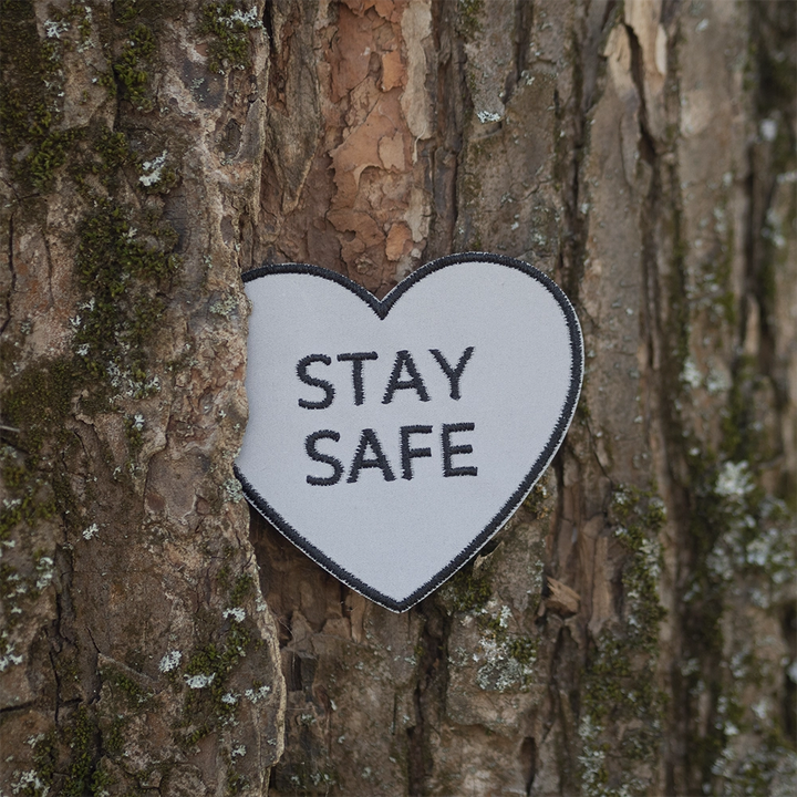 Brodeerattu heijastin haalarimerkki, sydämen muotoinen, teksti: Stay safe. Taustalla puunrunko.